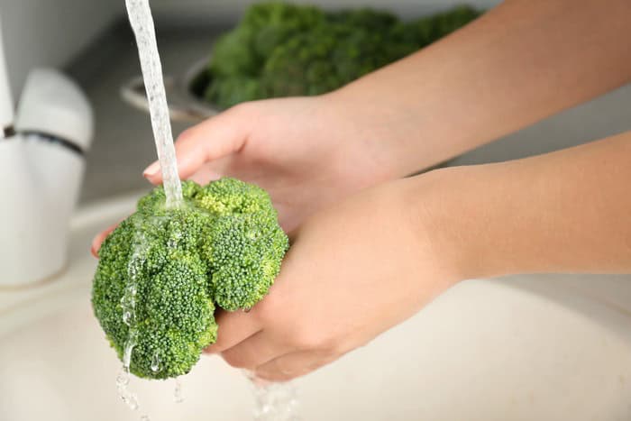 Manos lavando ramilletes de brócoli bajo el agua antes de cocinar brócoli con bechamel